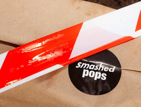 Verpackung mit rot-weißem Absperrband-Motiv und Smashed-Pops-Aufkleber